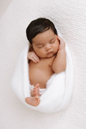 A sleeping newborn baby wrapped in white swaddling cloth rests peacefully in a curled position.