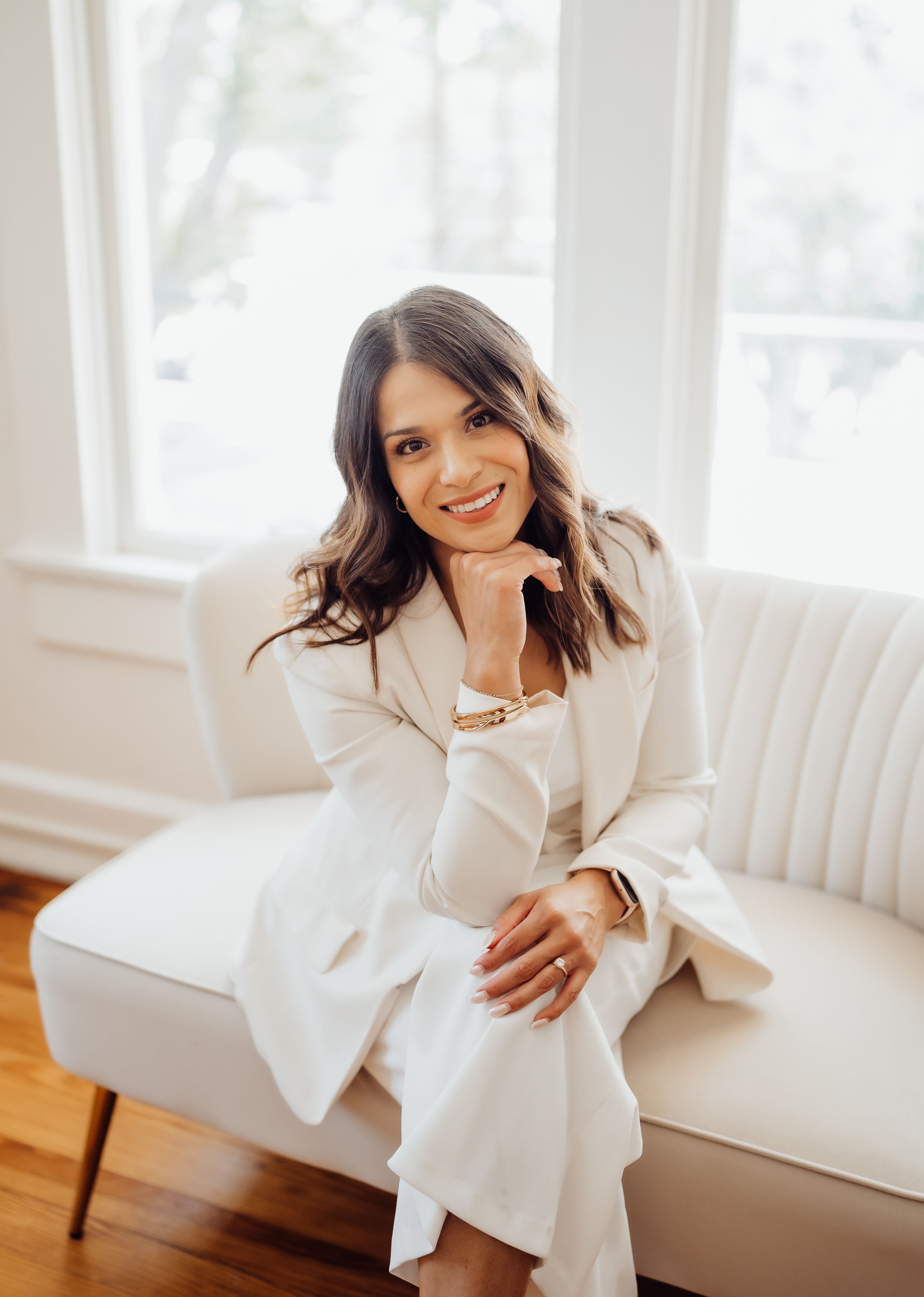 Woman in white business suit sitting elegantly on modern couch near bright windows.