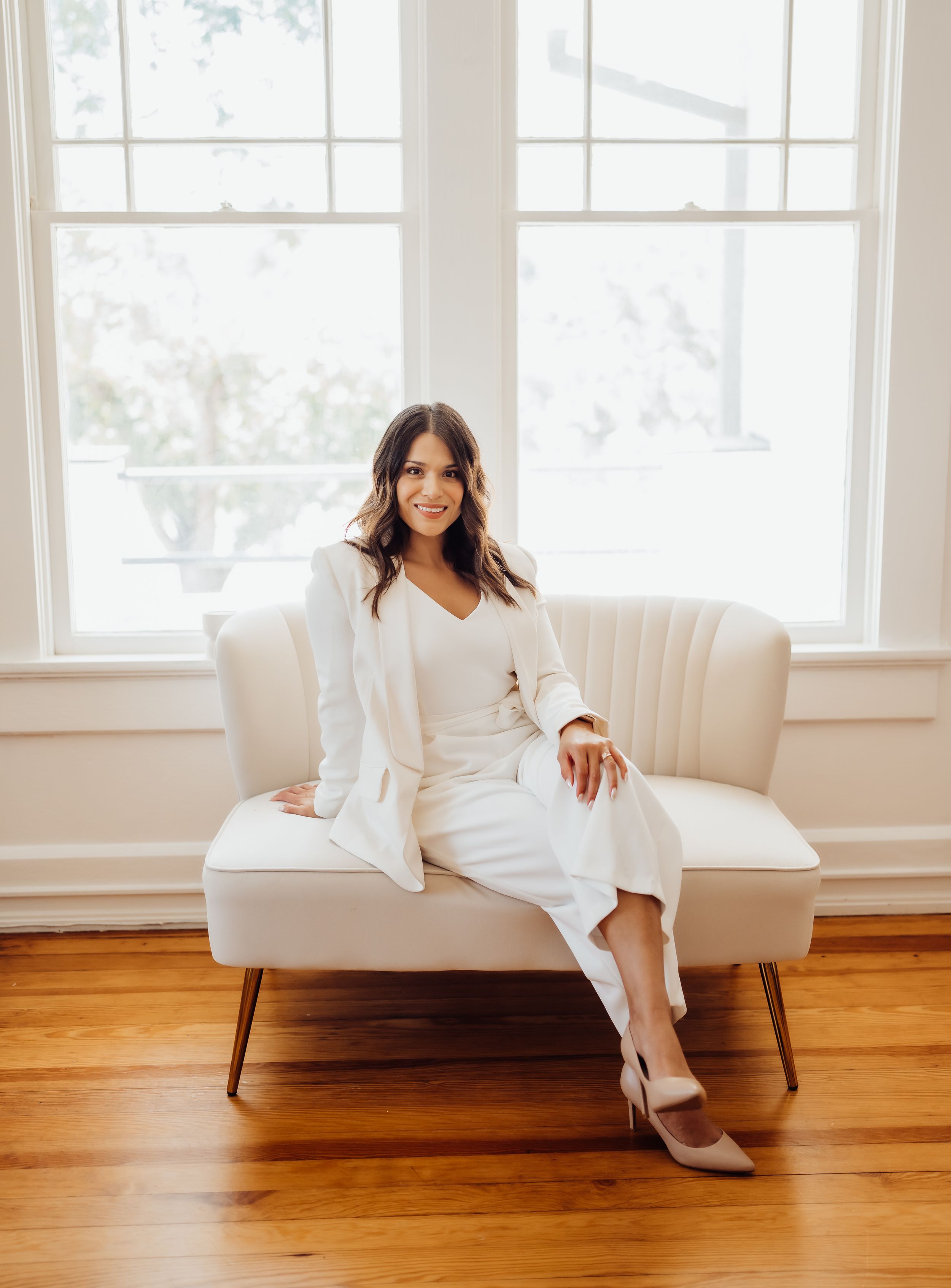 Professional woman in all-white outfit poses confidently on vintage-style settee.