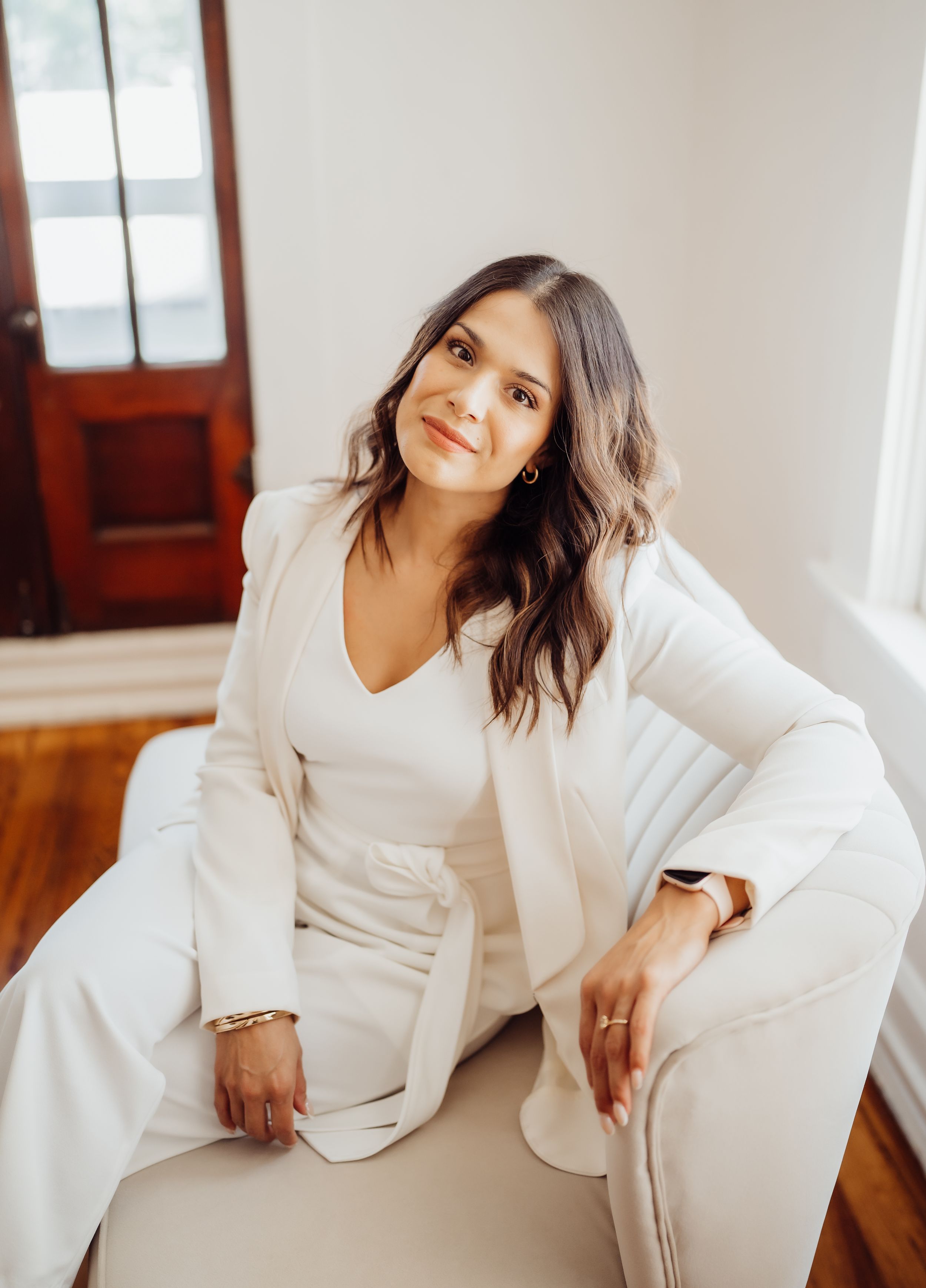 Professional portrait in all-white outfit sitting comfortably on cream colored chair in bright interior setting.