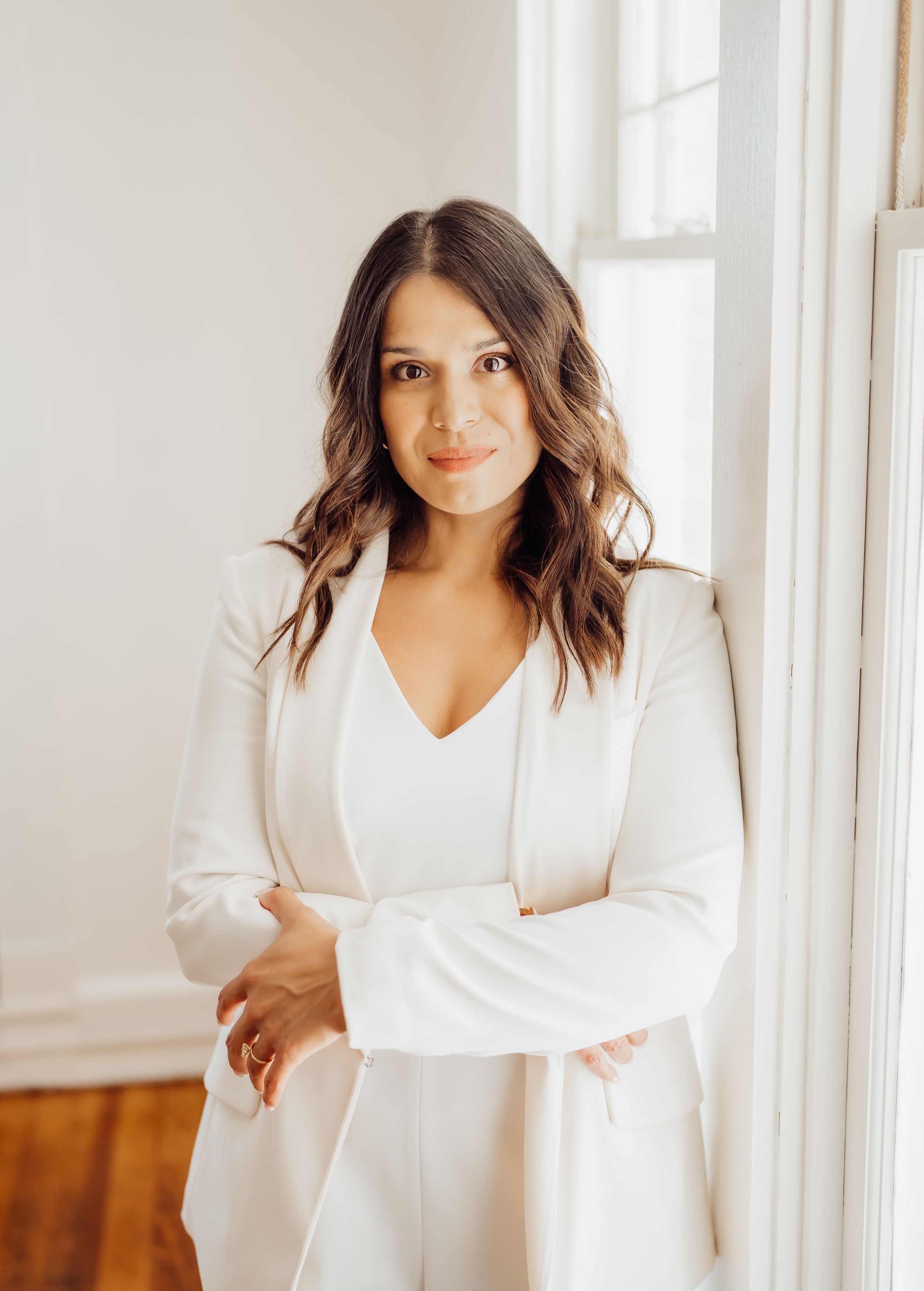 Professional headshot against white wall wearing crisp white blazer and top with wavy hair.