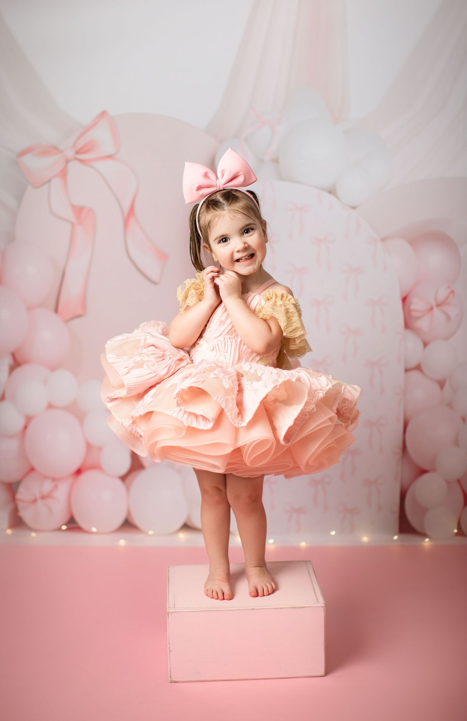 A toddler in a ruffled pink dress poses on a pedestal against a dreamy peach backdrop with floral decorations.