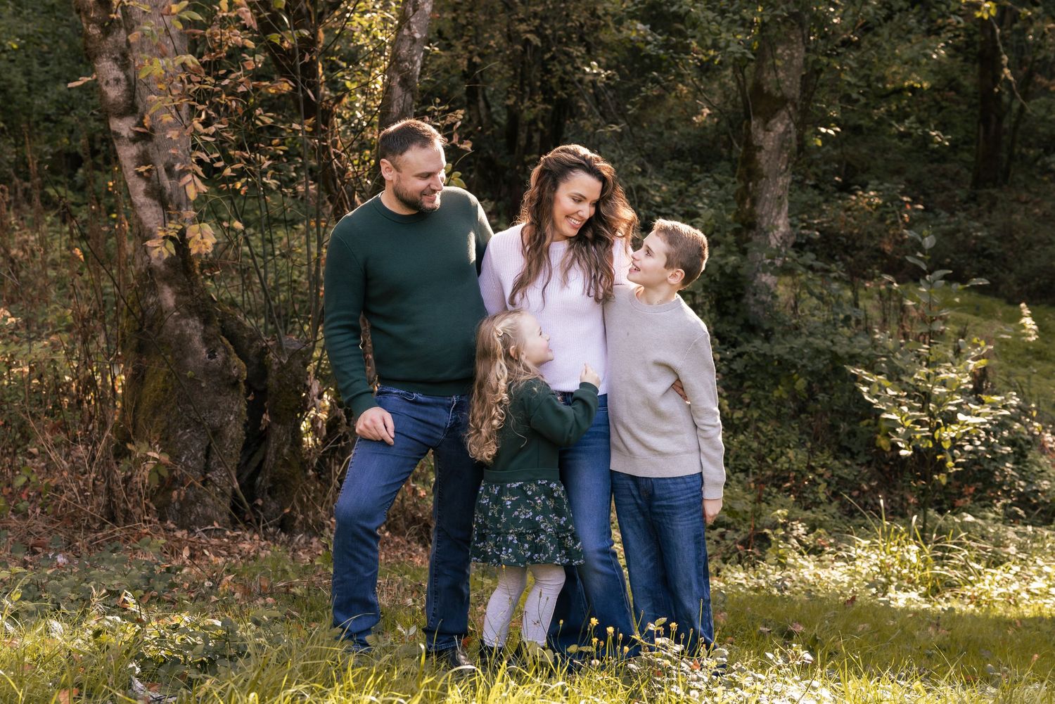 A family stands together outdoors in casual wear against a wooded backdrop on a sunny fall day.