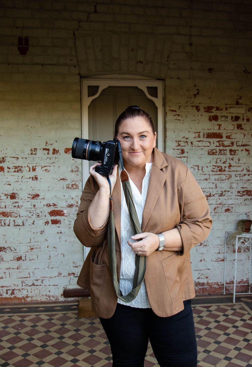 Professional photographer standing with camera in front of distressed brick wall wearing tan blazer and striped shirt.