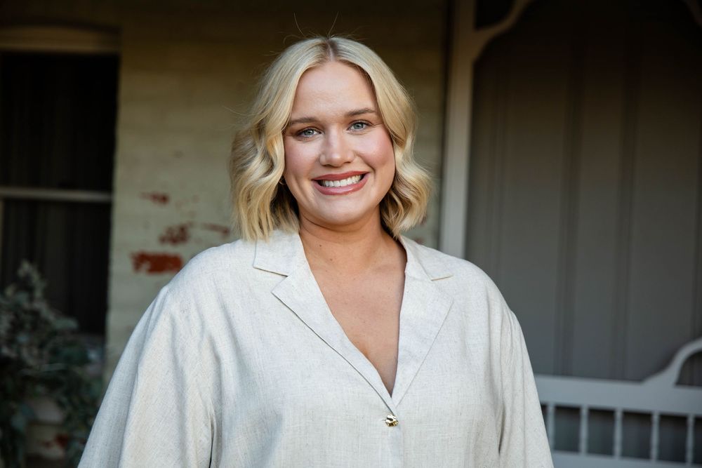 A person wearing a white button-down top stands against a dark background while smiling warmly at the camera.