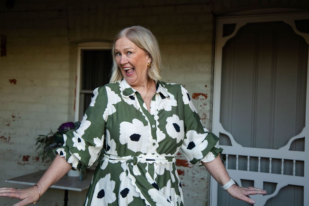 A person in a green and white floral dress stands against a white house exterior with a casual, relaxed pose.