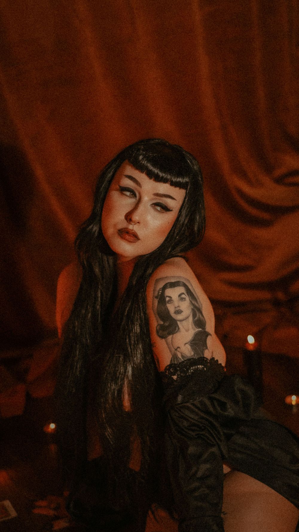 A gothic boudoir photography with red velvet curtain backdrop showing a vintage tattoo sleeve in dramatic lighting.