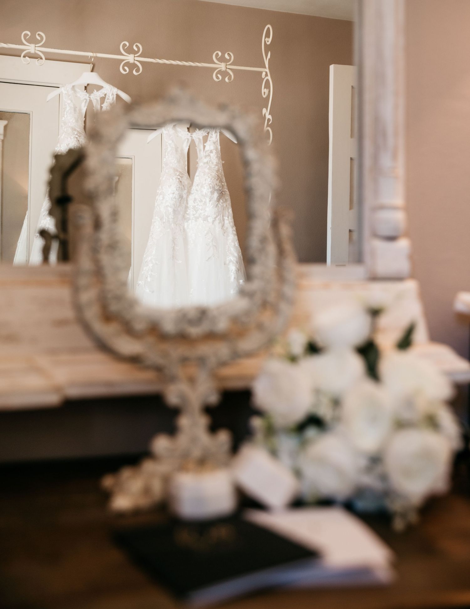 White wedding dress hanging on ornate wall mount with floral arrangements and vintage mirror.