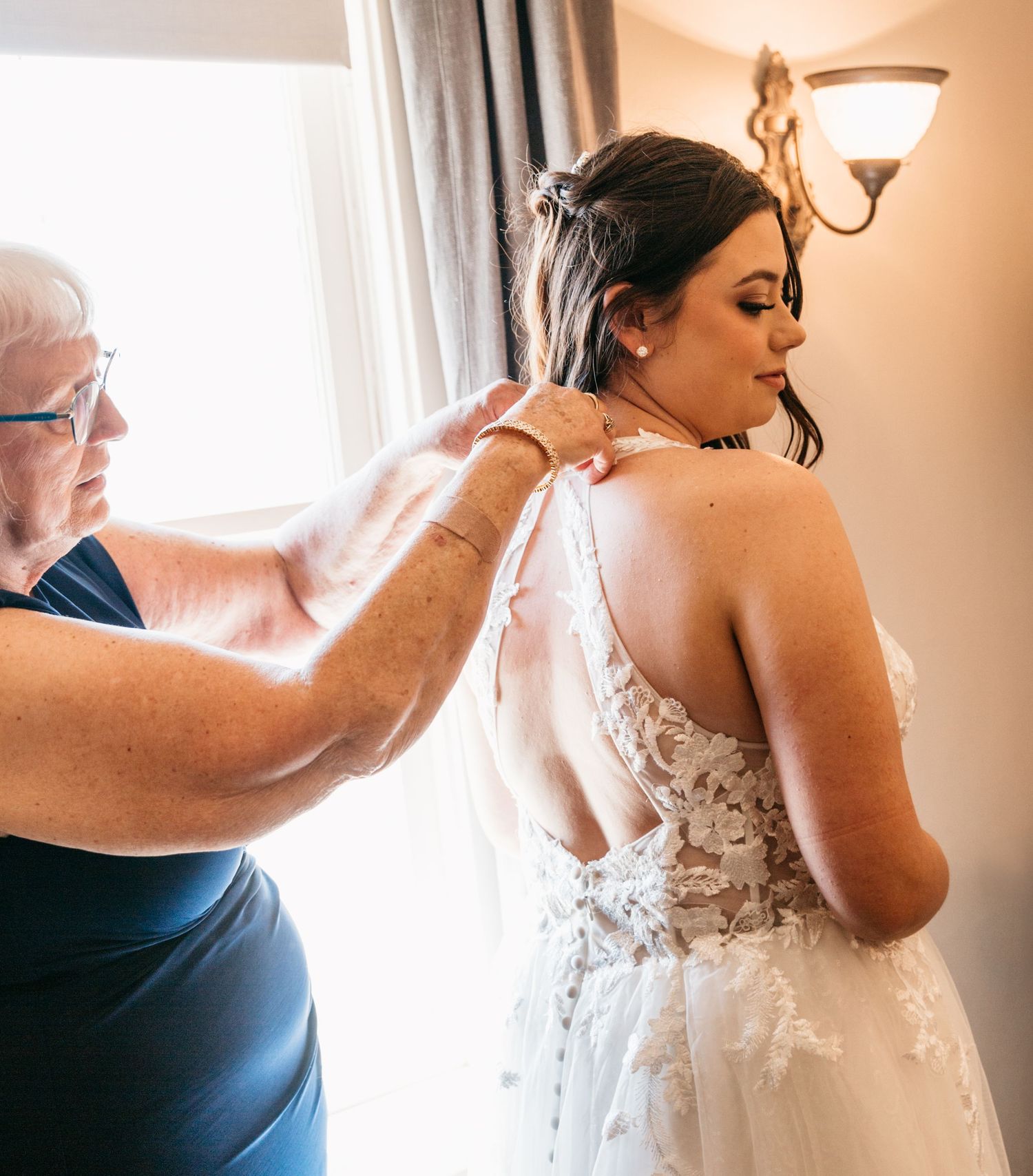 Helper assisting with bride's dress preparation in warmly lit room.