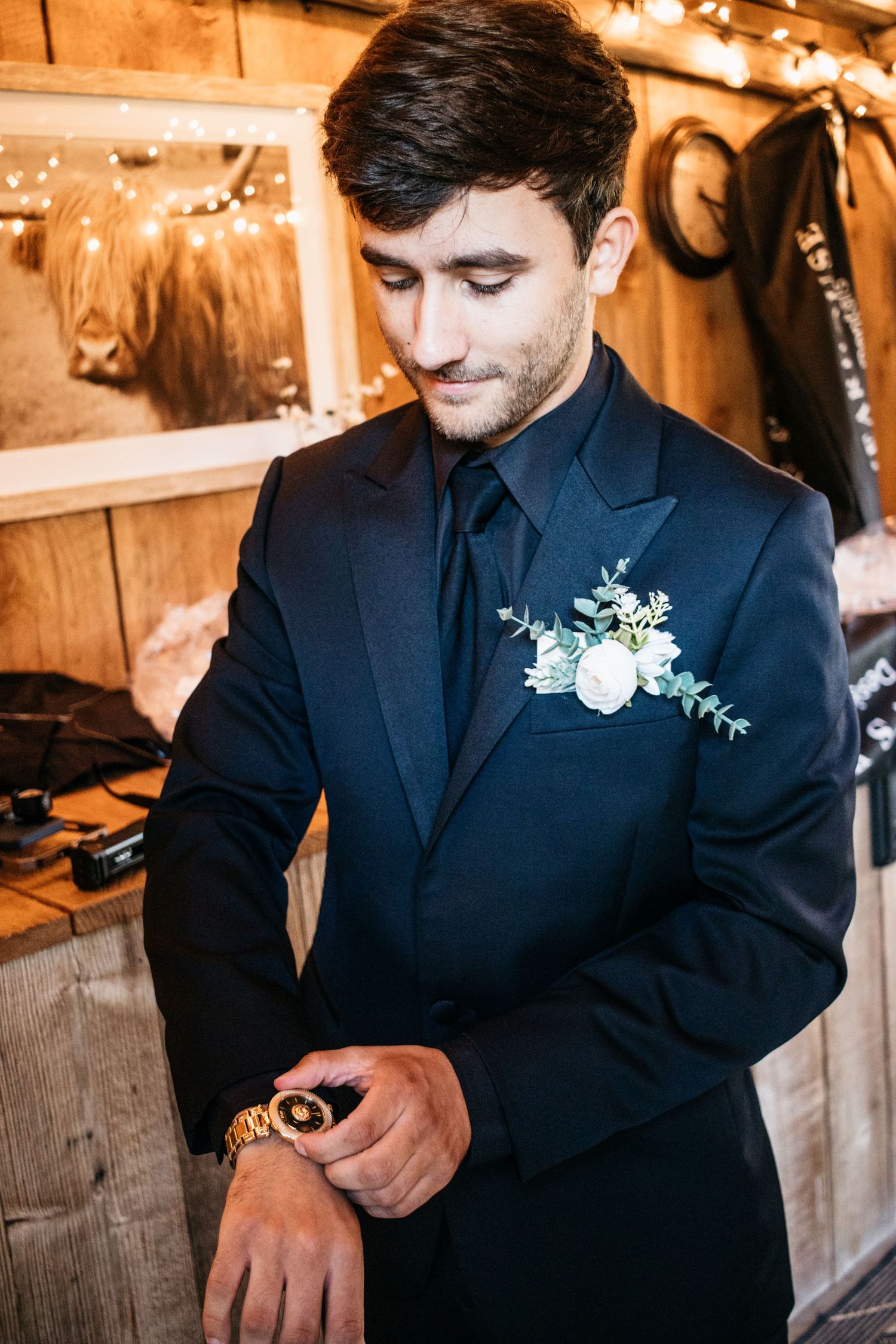 Groom in navy suit with white boutonniere checking time in rustic getting ready room.