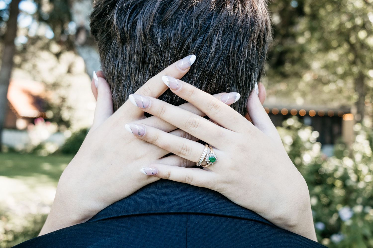 Hands wearing an emerald engagement ring rest on a shoulder outdoors.