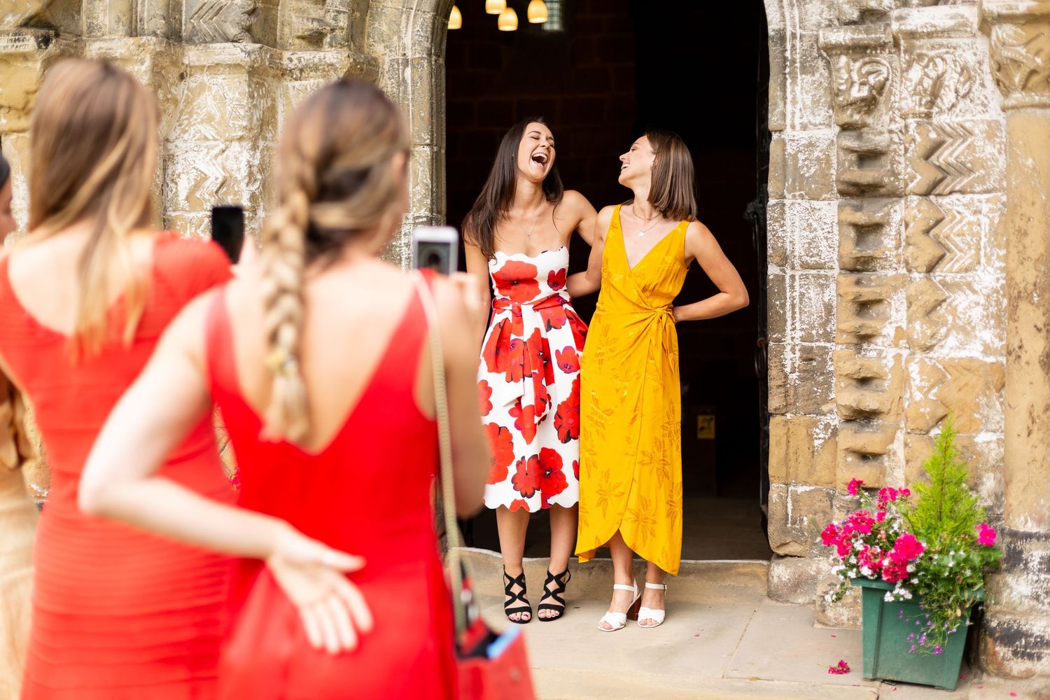 Group of women in bright dresses pose and laugh outside historic stone building.