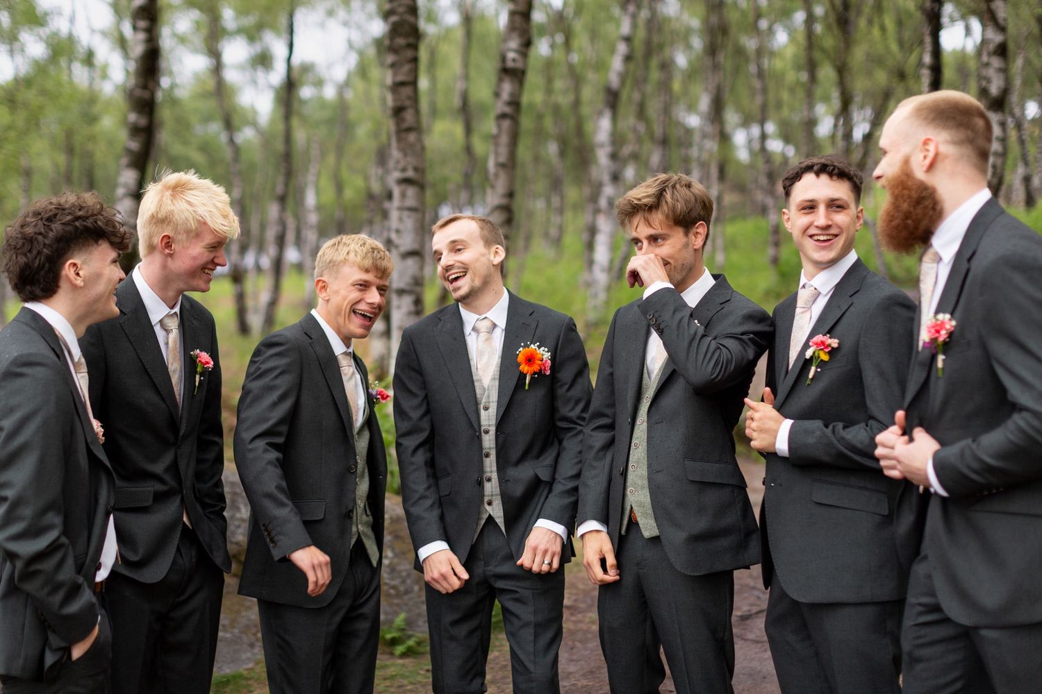 A group of groomsmen in dark suits share a candid laughing moment together in a wooded outdoor setting.