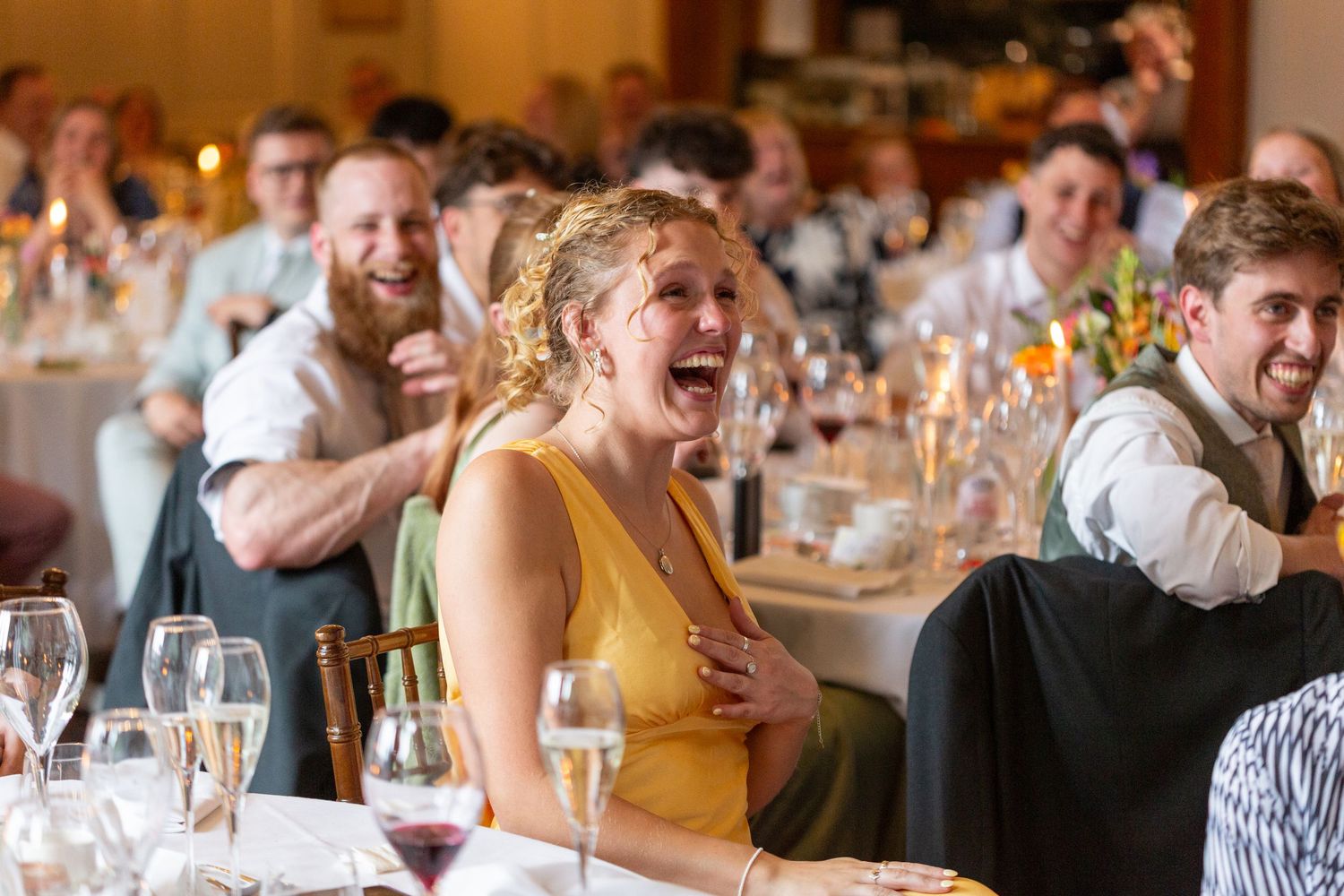 Happy wedding guests are seen smiling and laughing during an emotional reception speech at a brightly lit venue.