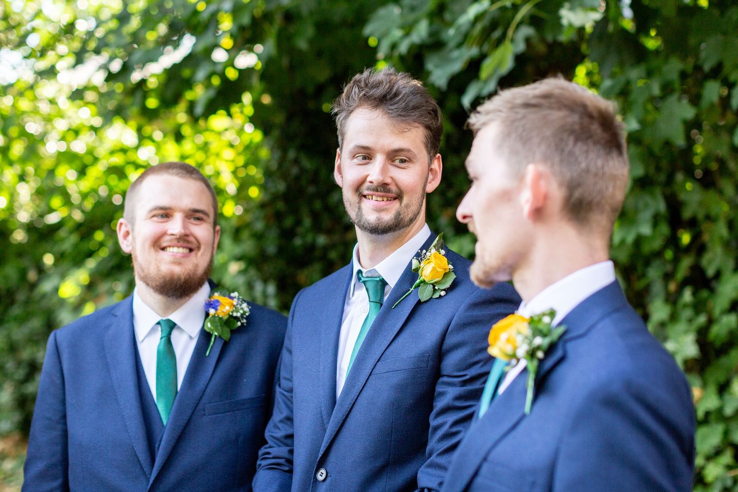 Three groomsmen wearing navy suits with emerald ties share a candid moment outdoors.