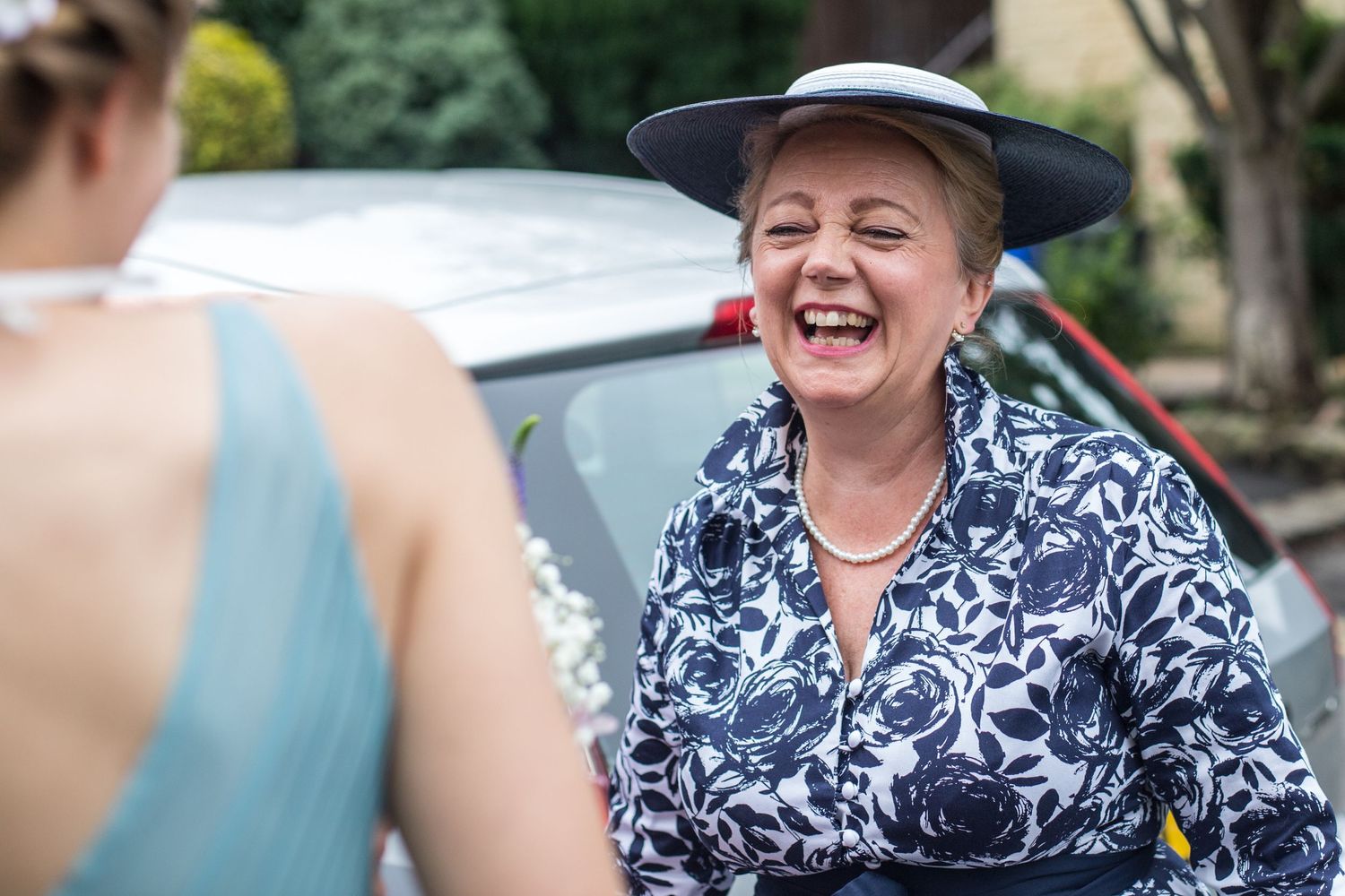 Guest in floral print dress and hat shares a hearty laugh during outdoor celebration.