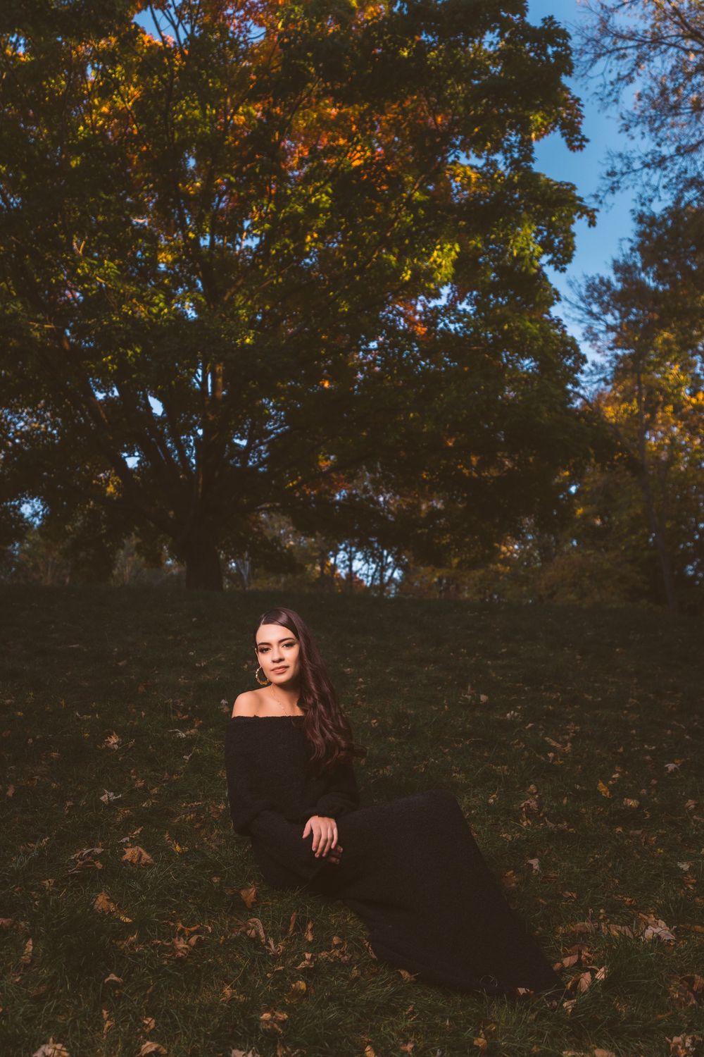 A person in black attire poses among autumn trees with golden foliage.
