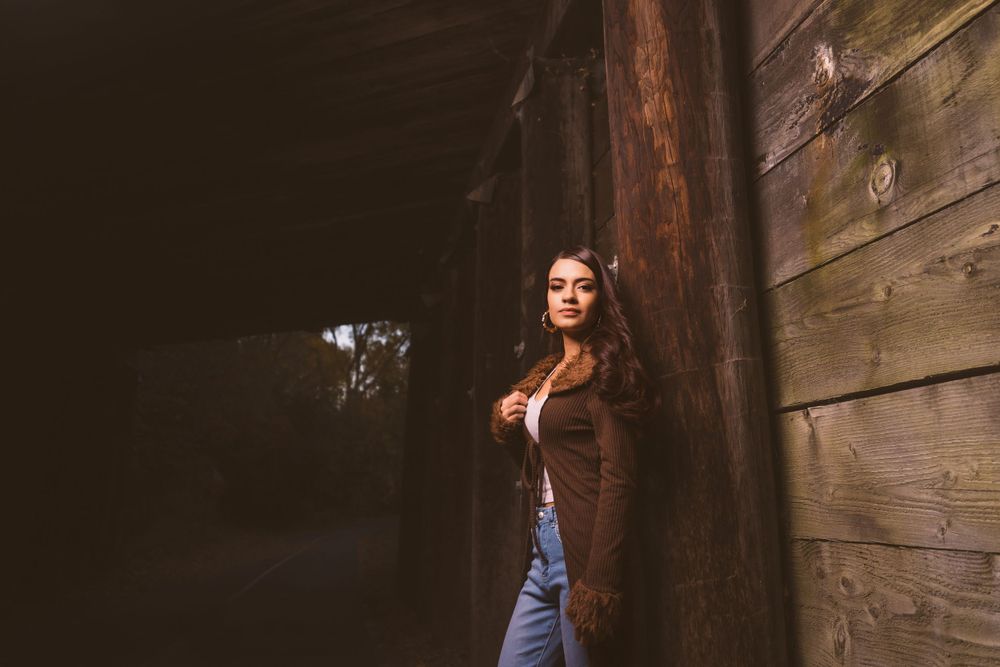 A figure poses against rustic wooden walls in moody lighting.