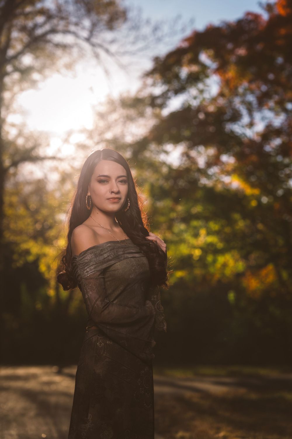 A figure in dark lace poses outdoors during golden hour with autumn trees in the background.