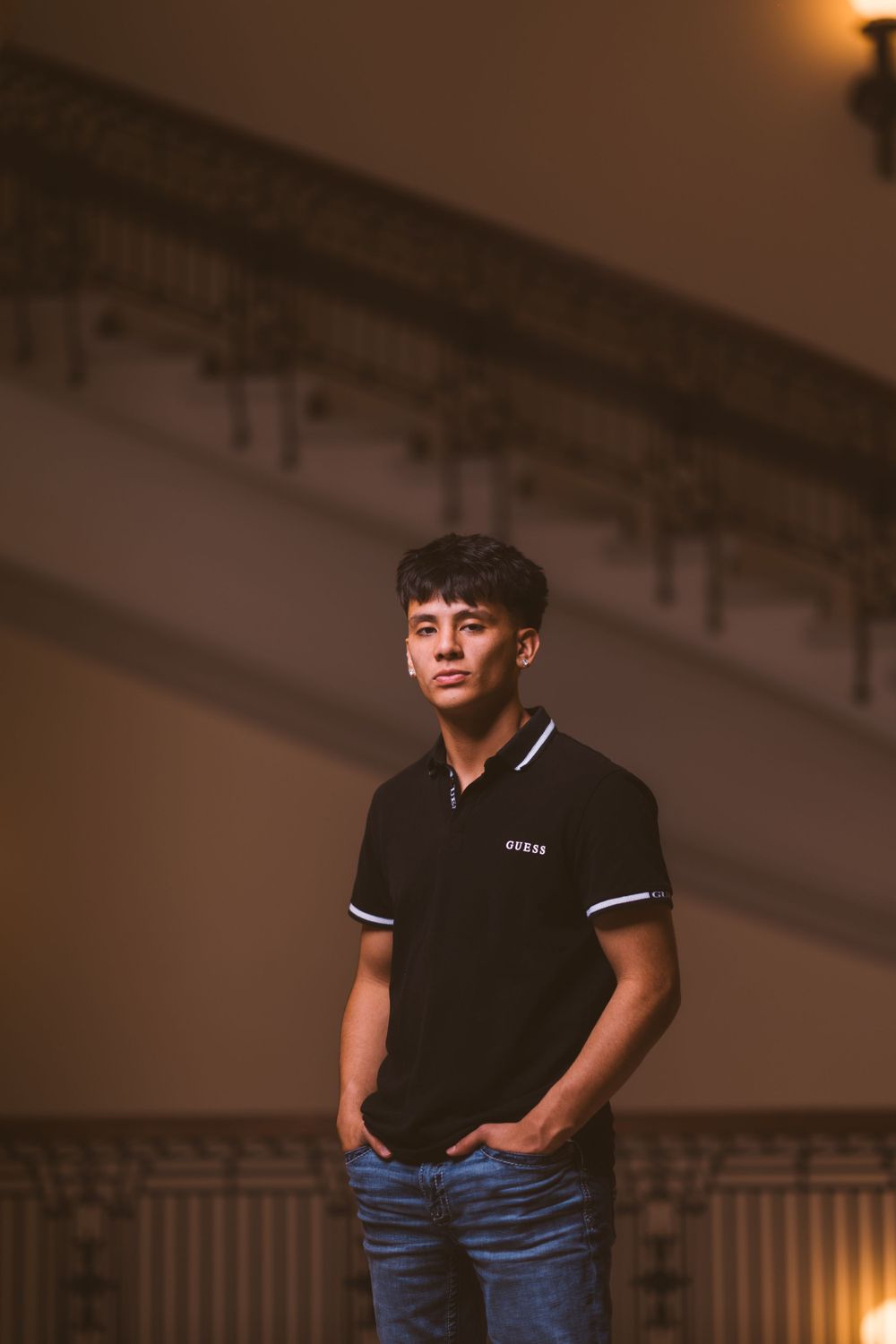 A person in a black polo shirt and jeans poses casually in an indoor setting with warm lighting.