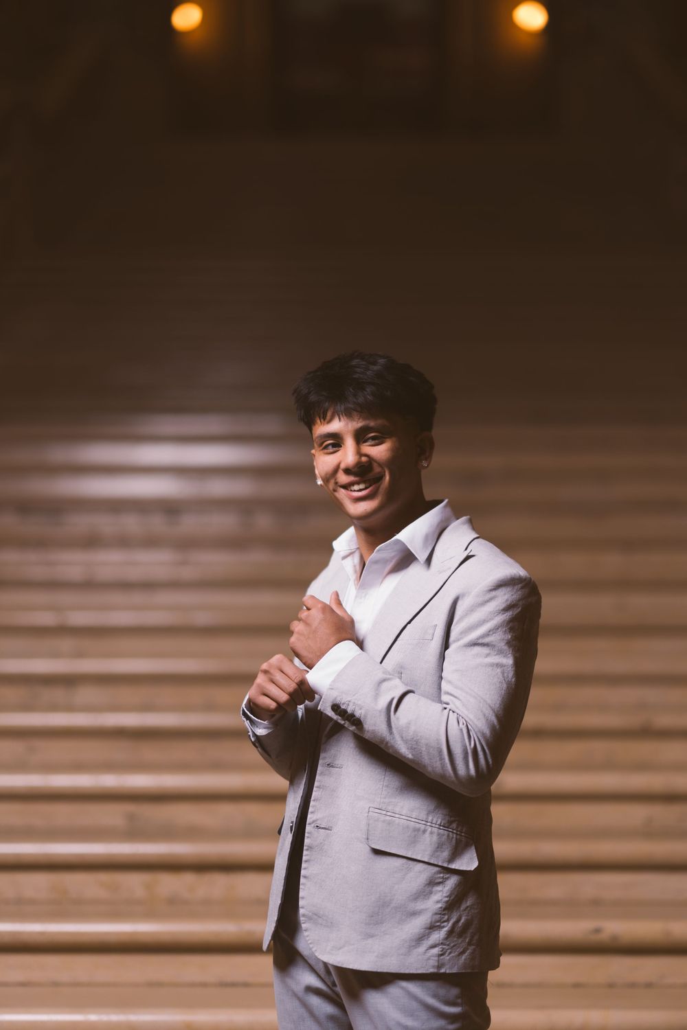A figure in a light gray suit jacket poses confidently against a wooden backdrop.