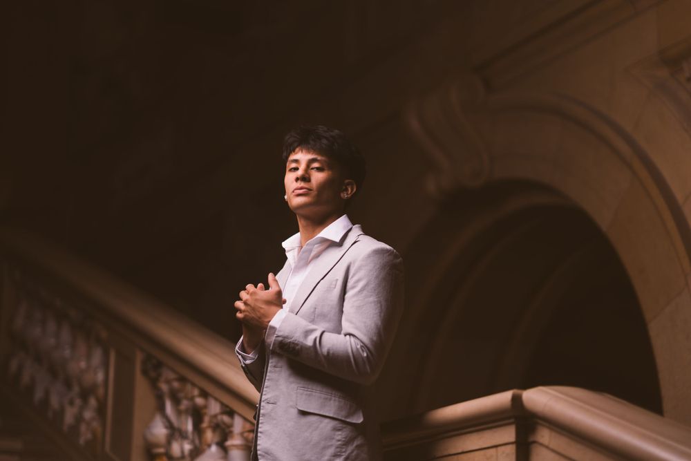 A figure poses expressively in a light colored suit against a dark architectural background.