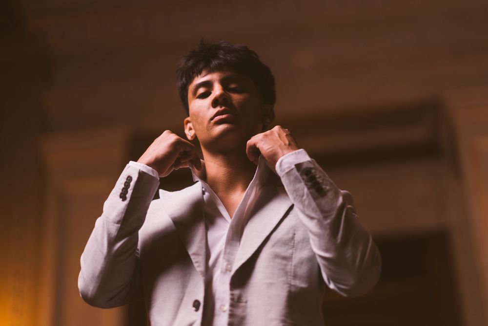 A person in a light gray blazer poses dramatically in an indoor setting with warm lighting.