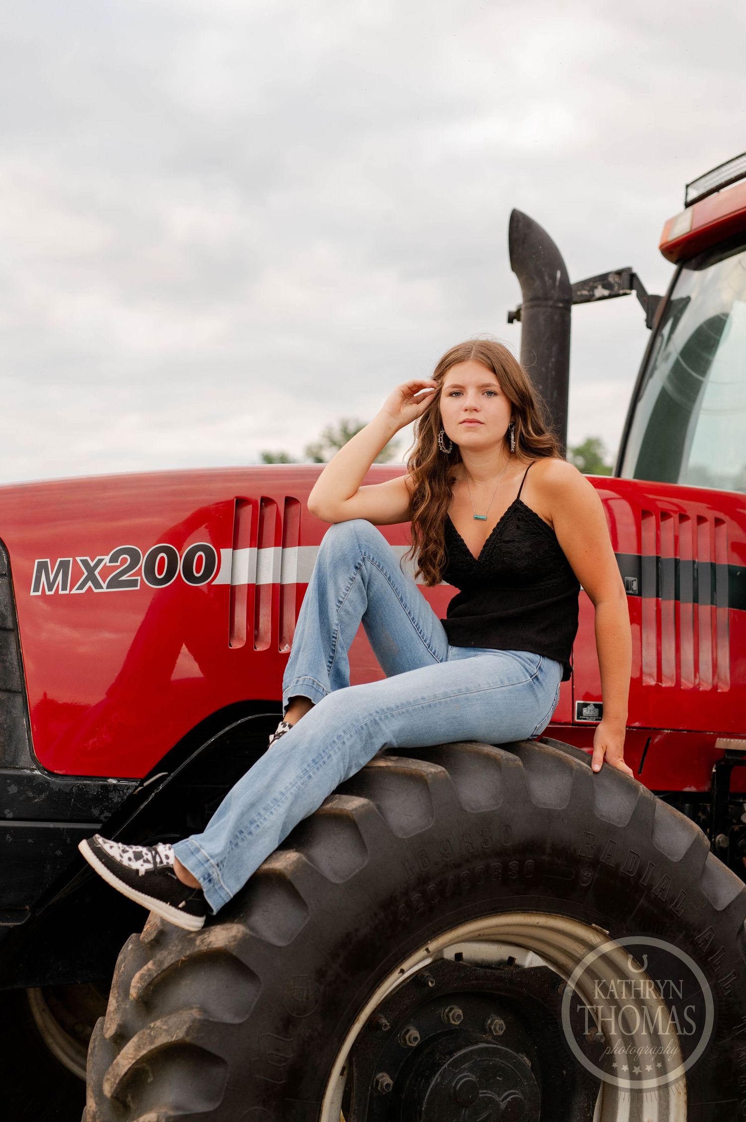 Someone poses casually on the large tire of a red MX200 tractor against a cloudy sky.