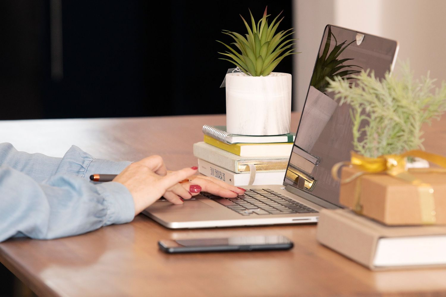 Personne travaillant au bureau avec un ordinateur portable, des livres et de petites plantes en pot dans un espace de travail moderne.