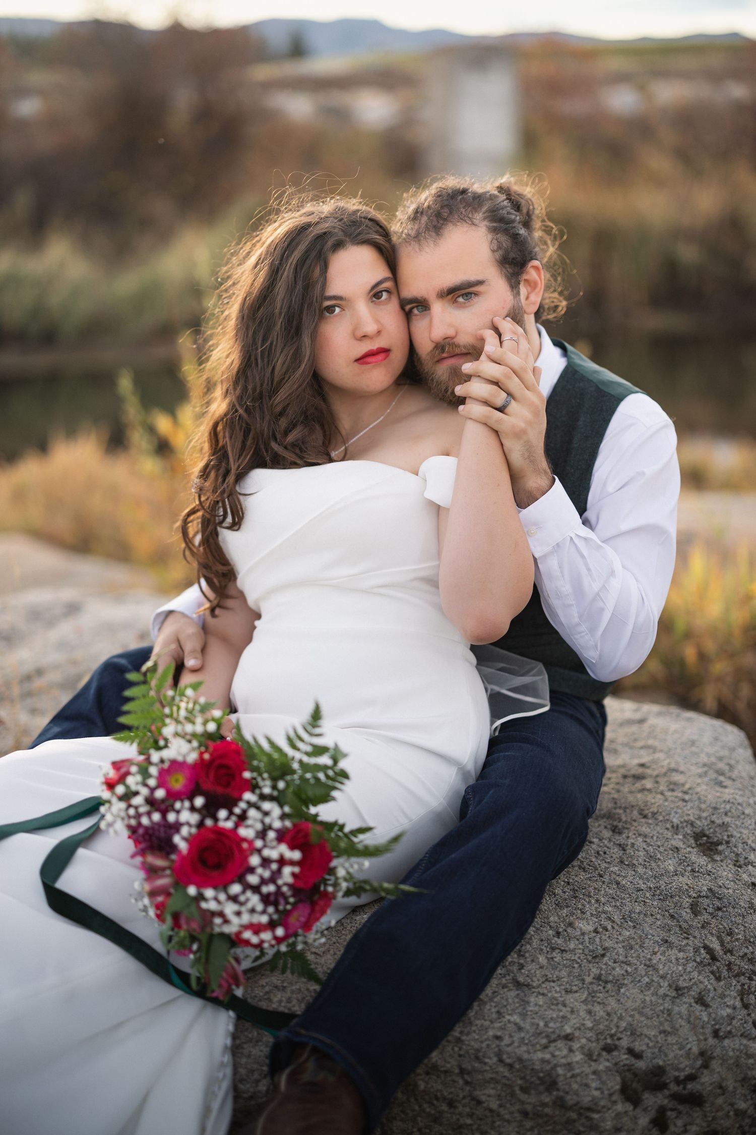 A romantic wedding portrait featuring a couple sitting on rocks with a red floral bouquet at sunset.
