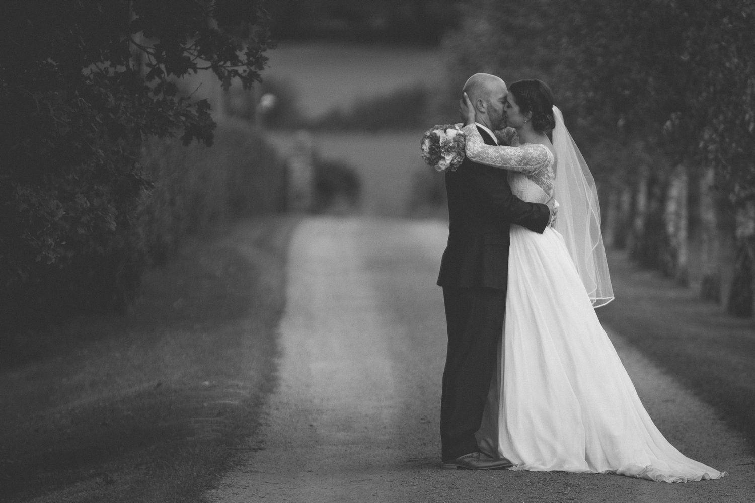 Editorial bridal portrait of a bride and groom in black and white embracing as the driveway fades into the distance. Stratton Court Barn