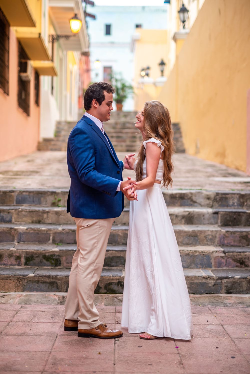 Couple holding hands on stone steps with colorful colonial architecture background.