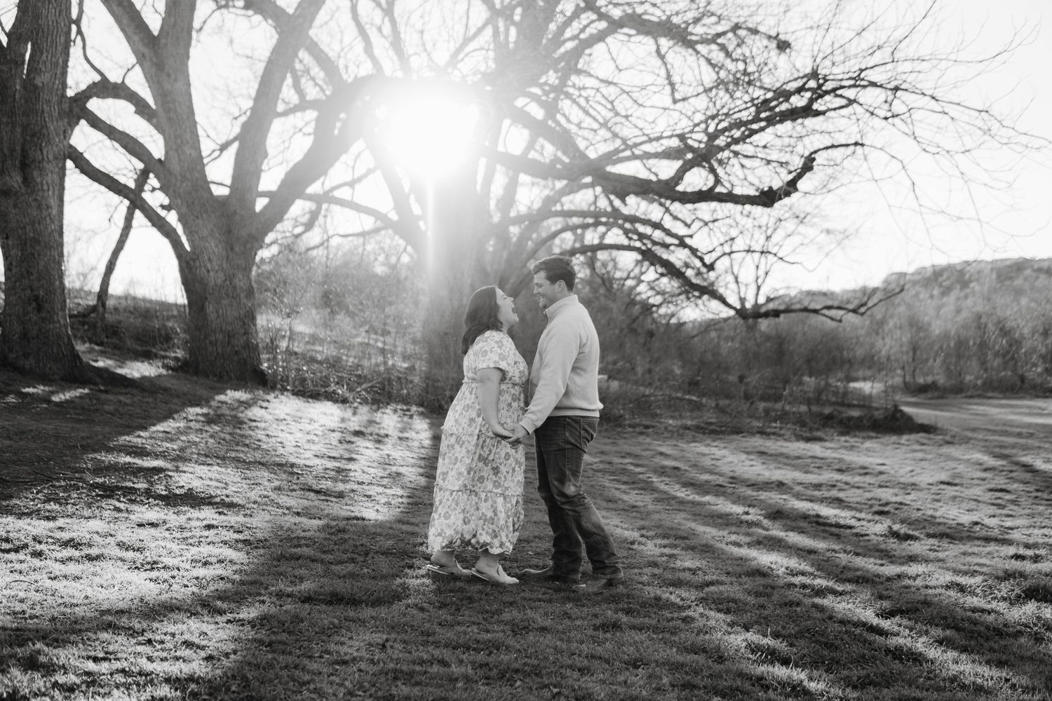 Black and white romantic photo of a couple embracing under a dramatic sunburst through bare winter trees.