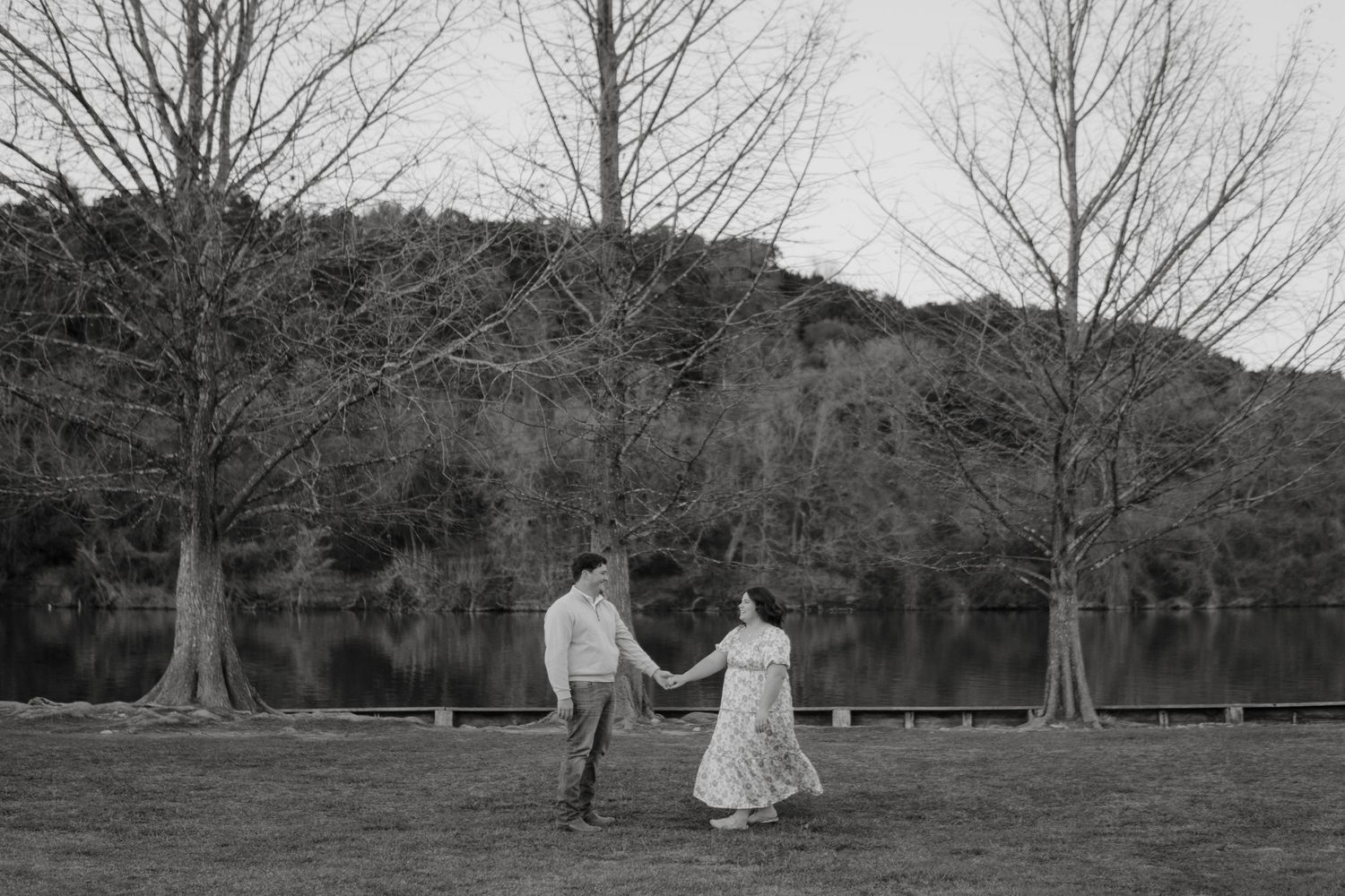 Two people in formal attire holding hands and walking across a grassy field with bare trees behind them in black and white.