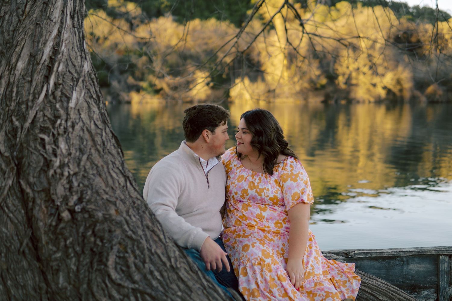 A couple shares a romantic moment while sitting by a lake with fall foliage reflecting in the calm water.