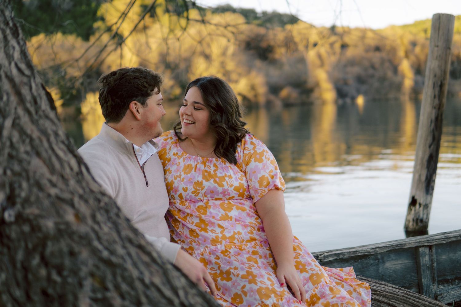 A couple shares a tender moment by a lake at sunset, with the woman wearing a floral dress.