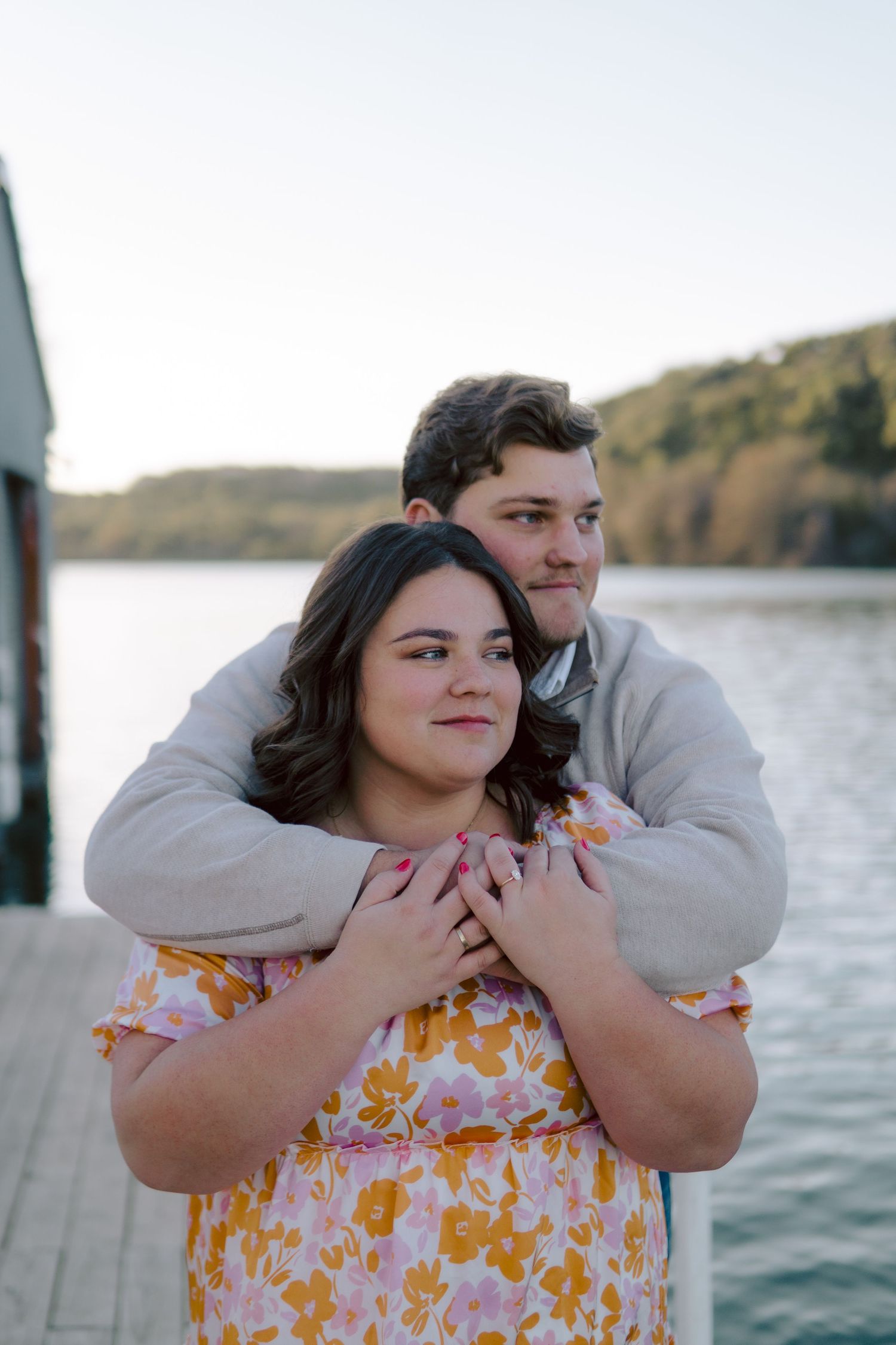 A couple embraces on a wooden dock by a lake at dusk, with the person in a yellow floral dress being held from behind.