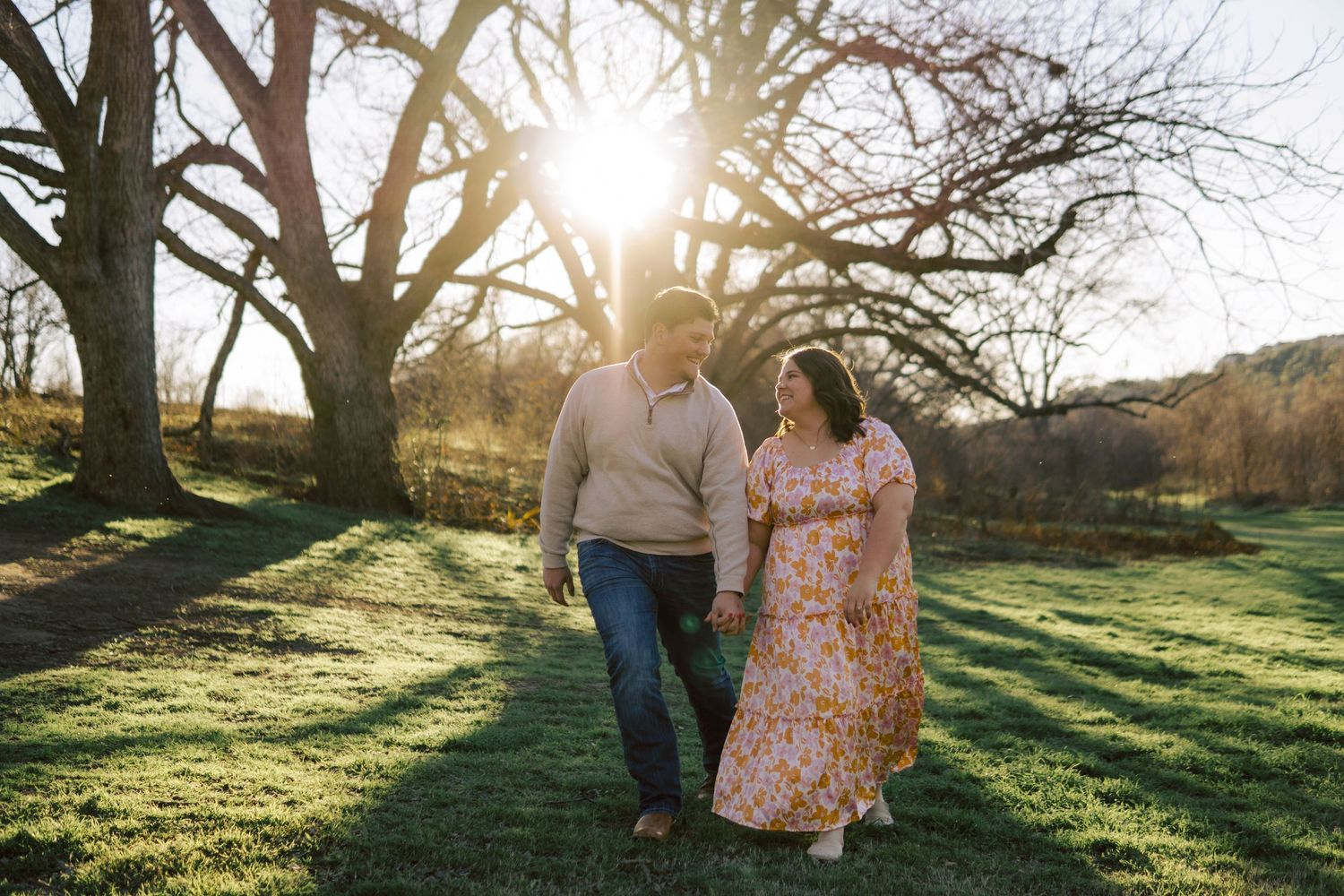 A couple walks together on a grassy hillside during golden hour sunlight filtering through bare trees in autumn.