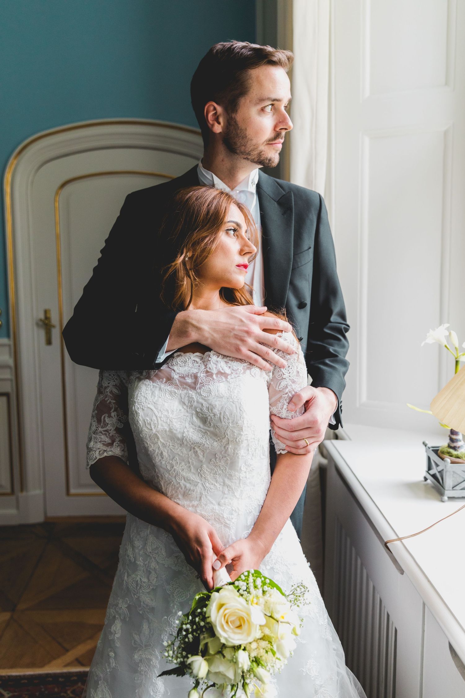 Romantic wedding portrait by window with couple in elegant attire holding white rose bouquet.