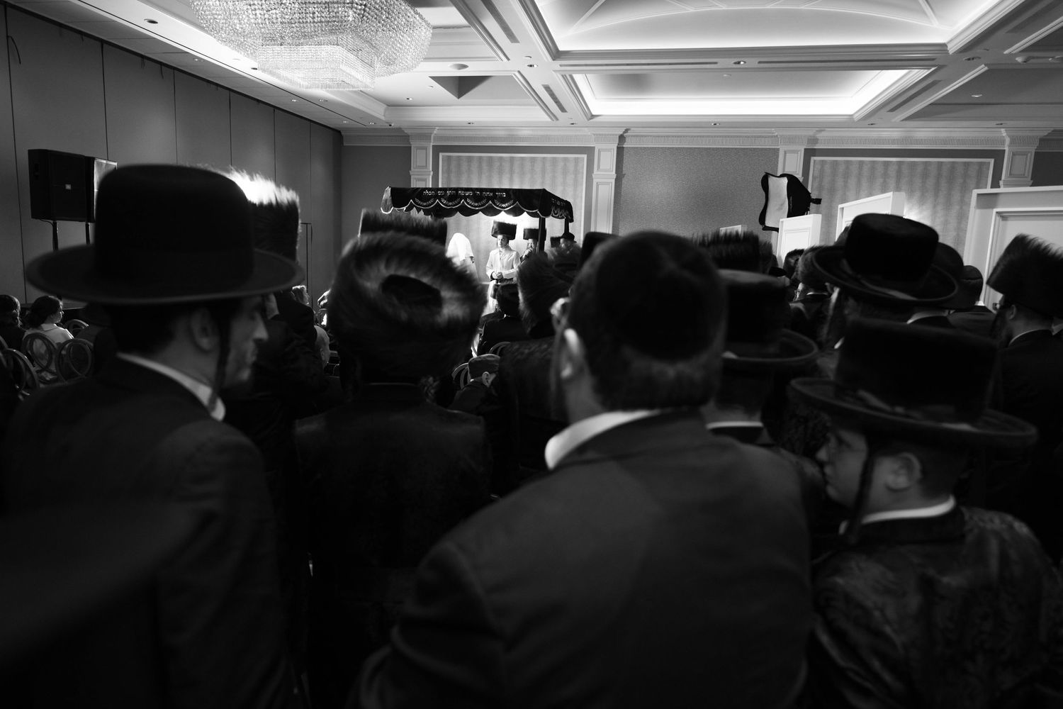 Black and white photo showing crowd of orthodox Jewish men in dark hats and suits gathered at an indoor event.