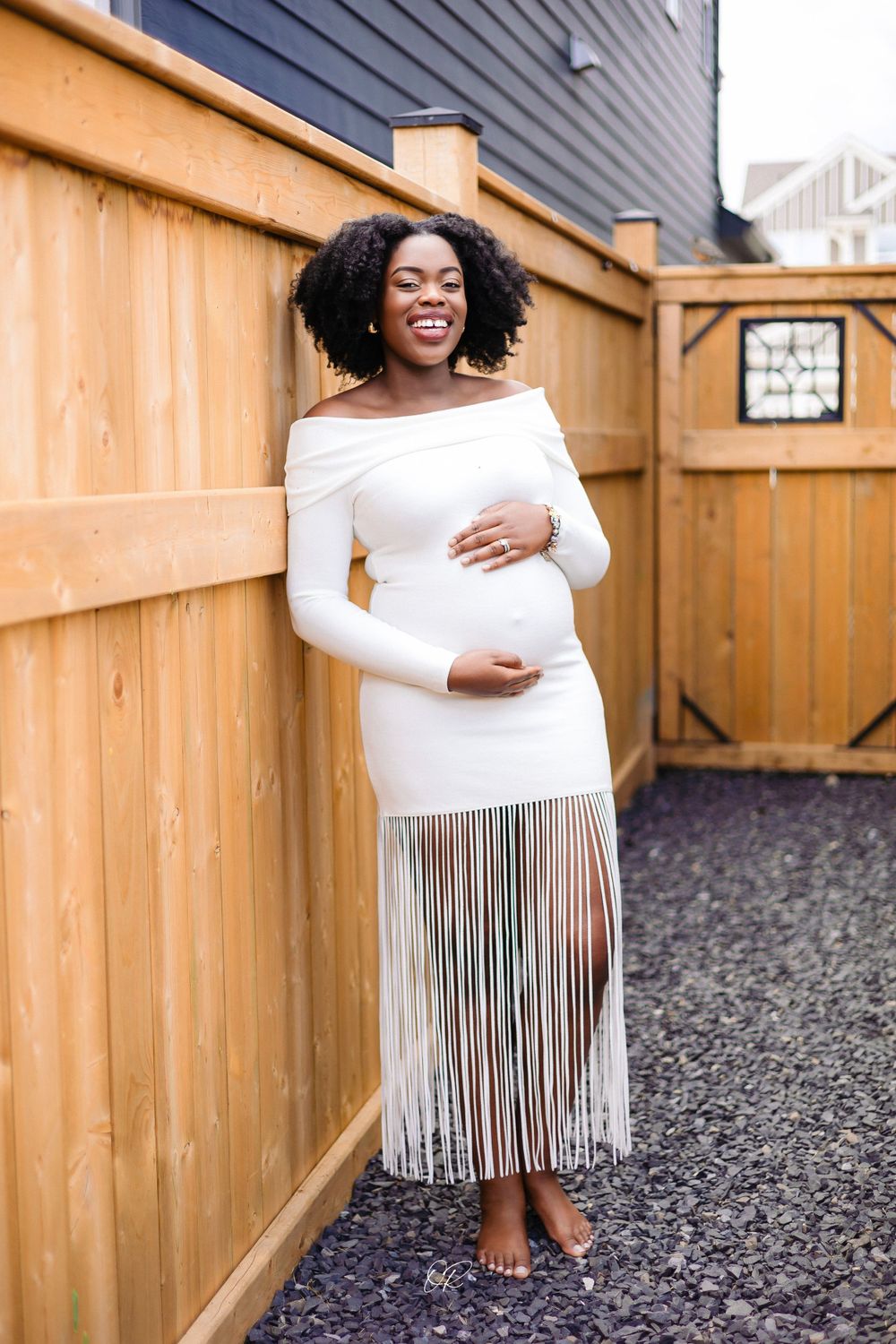 Maternity portrait in white fringe dress against wooden fence backdrop.