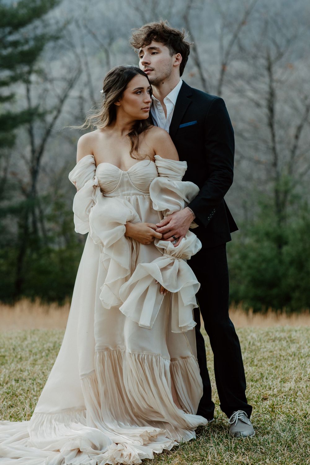 Outdoor wedding photo of couple with flowing off-shoulder dress and black suit against nature backdrop.