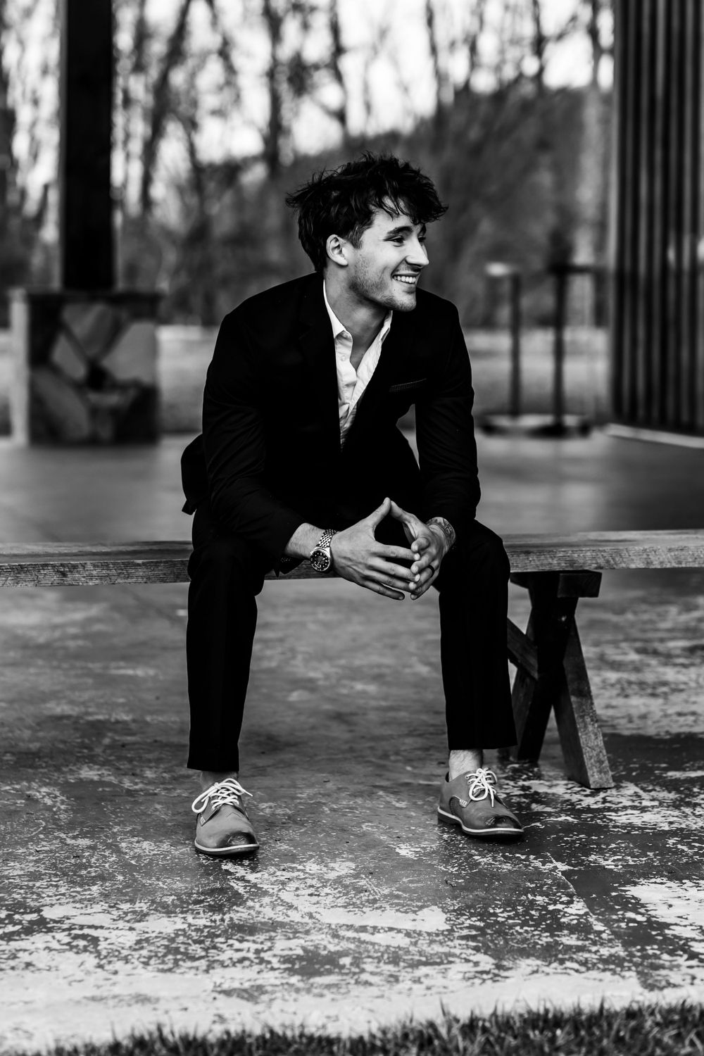 Black and white portrait of person in suit sitting on bench outdoors with joyful expression.