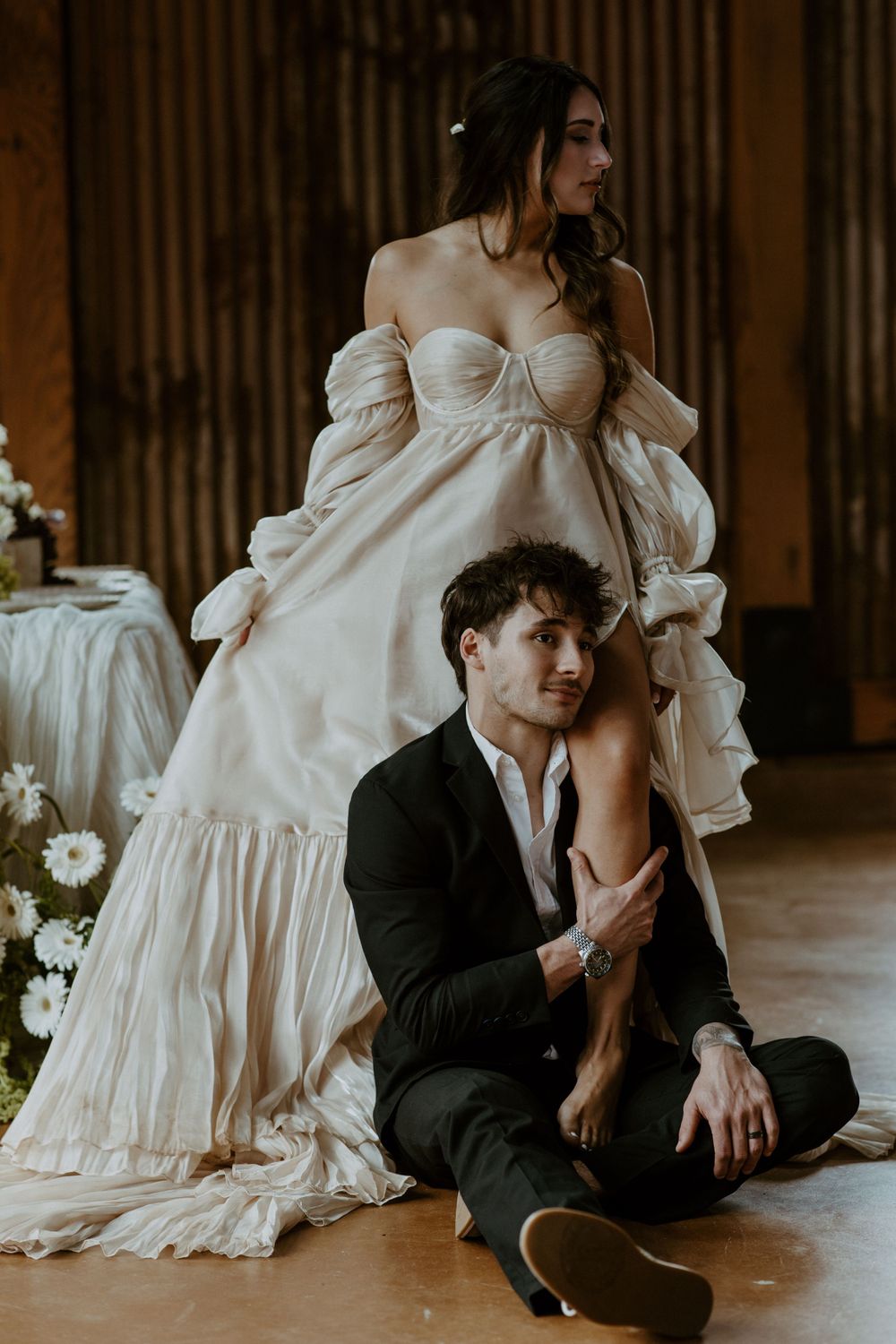 Romantic wedding portrait with couple in off-shoulder gown and dark suit sitting on rustic wooden floor for a styled shoot.