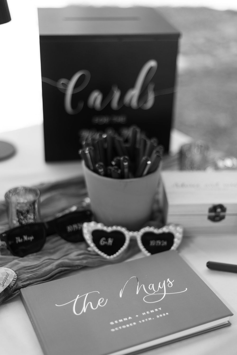 Black and white wedding detail shot of a card box, sunglasses, and guest book with calligraphy.
