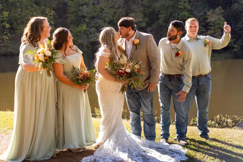 Wedding party poses by lakeside at sunset with bridesmaids in sage green dresses and groomsmen in casual attire.