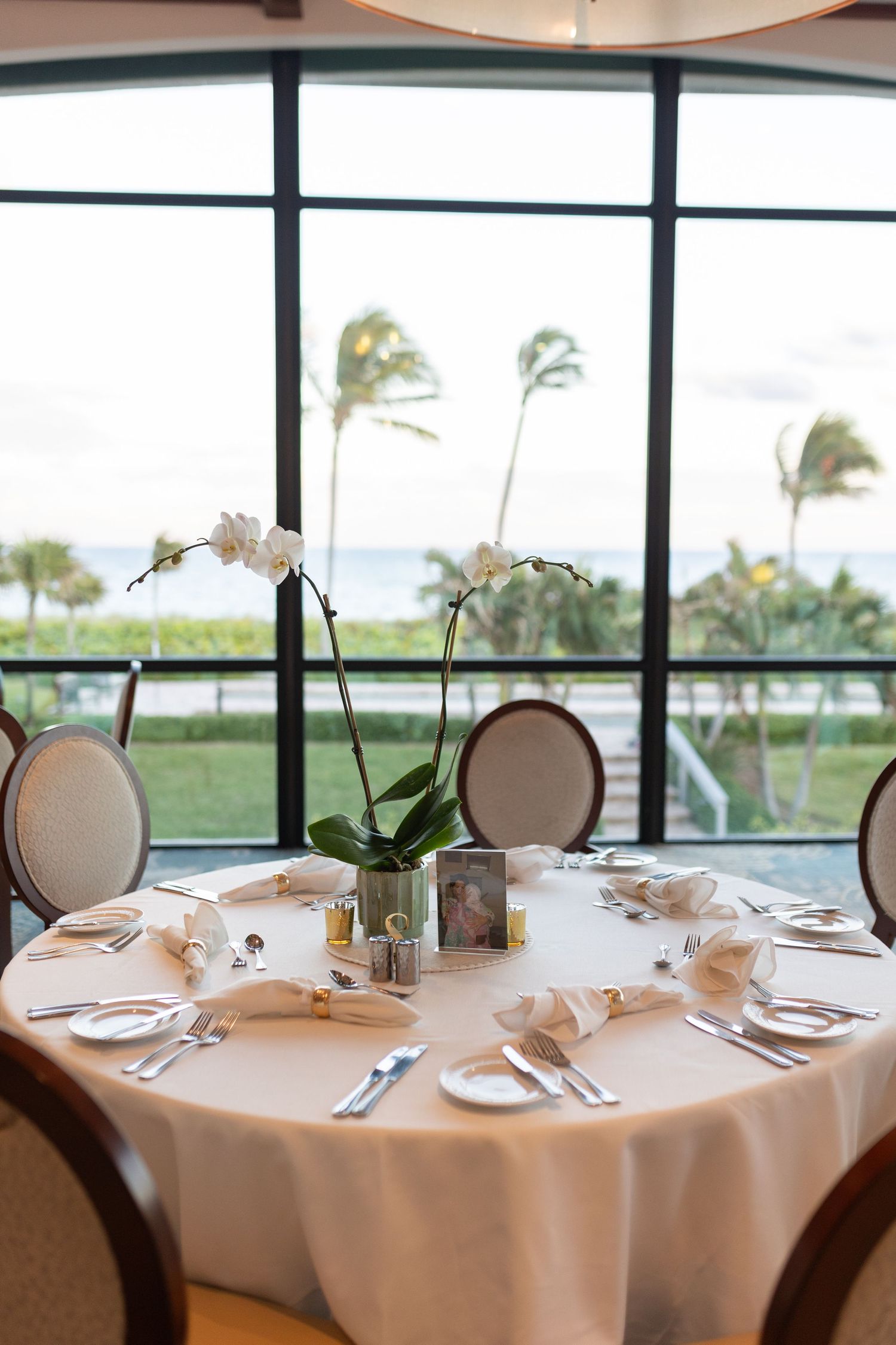Elegant beachfront restaurant table set with white orchid centerpiece and palm tree views.