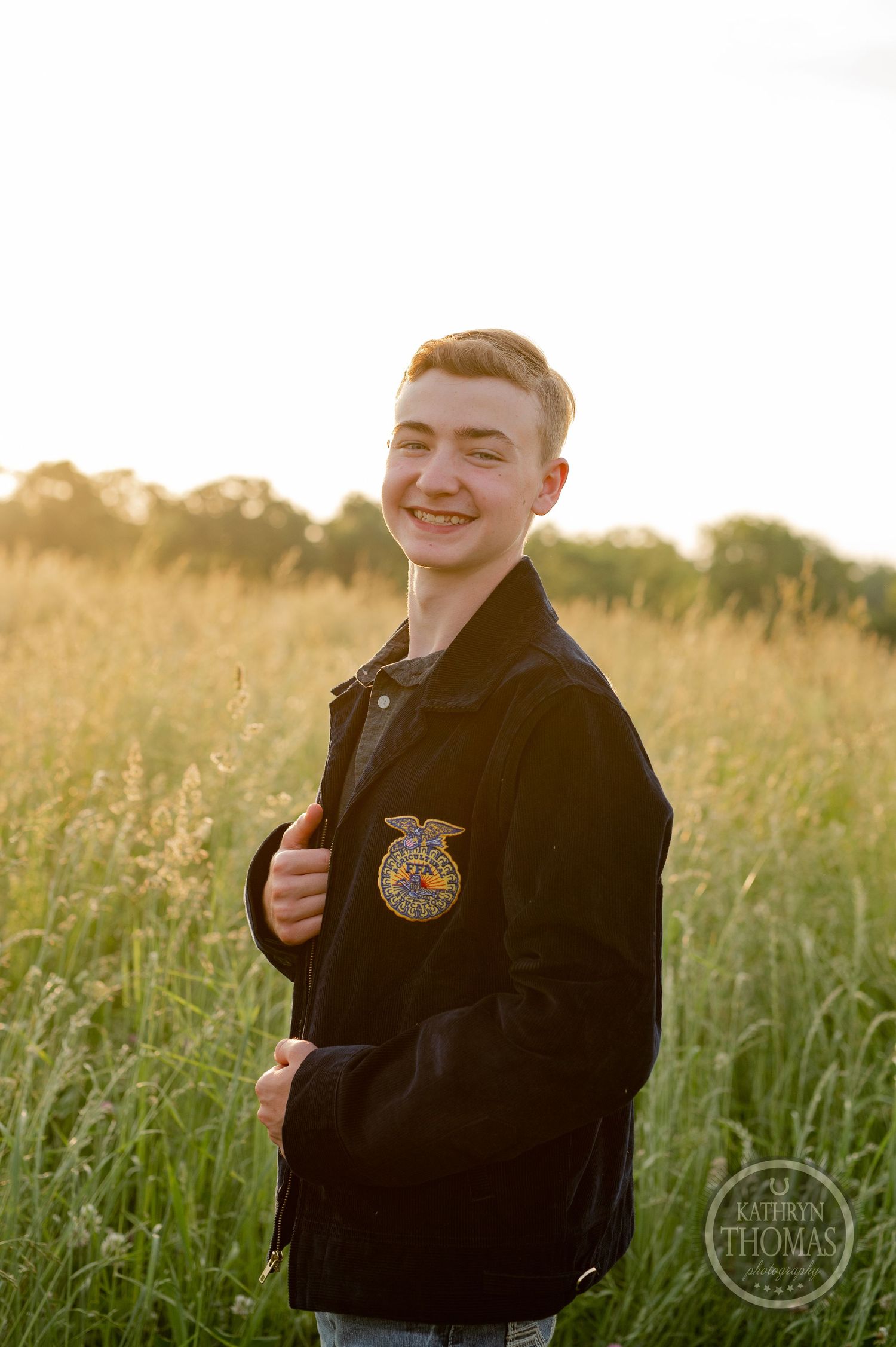Outdoor portrait in golden field showing FFA member wearing black jacket with emblem at sunset.
