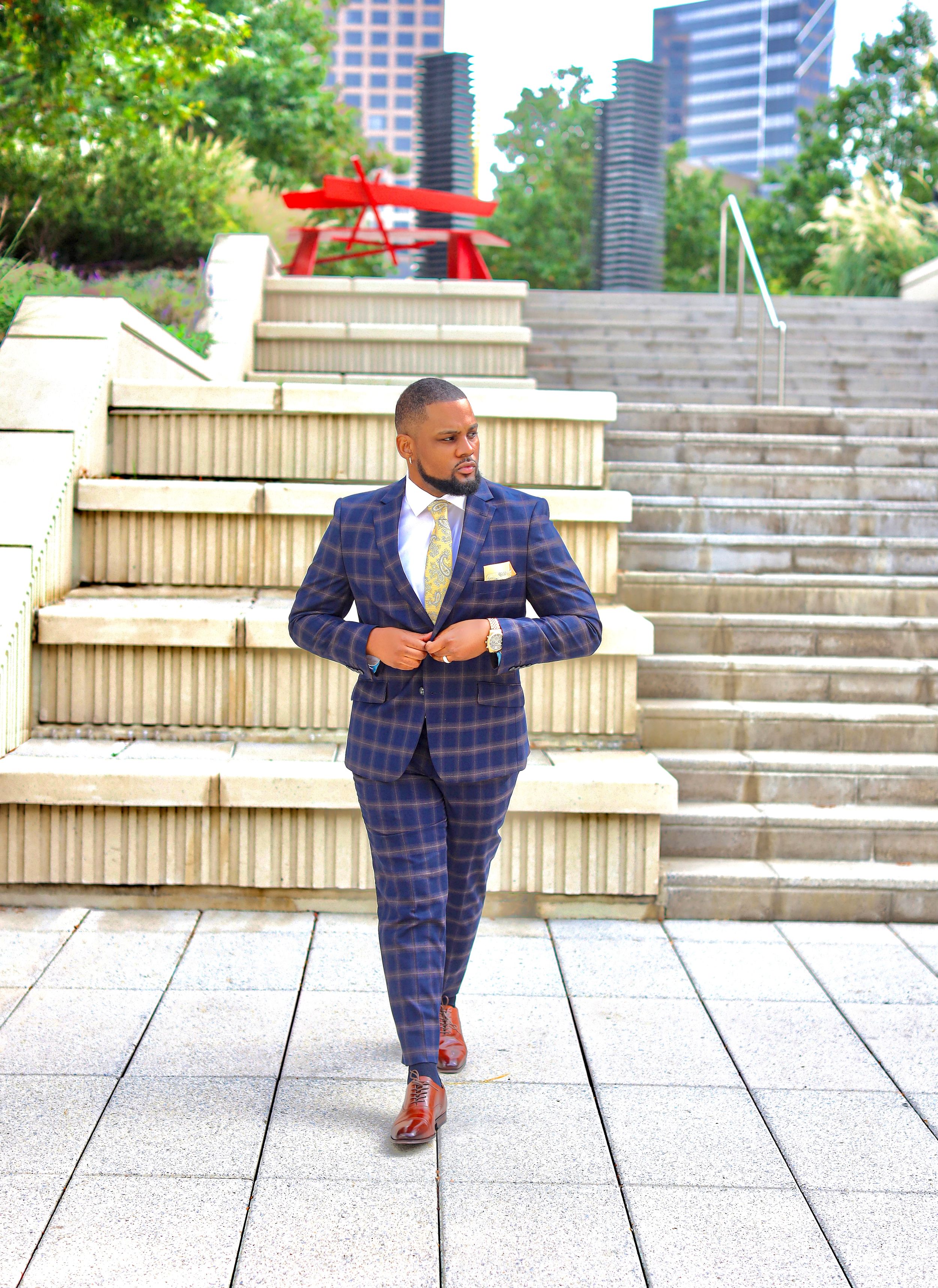 A person in a navy plaid suit and brown shoes poses confidently on concrete outdoor steps with city buildings in background.