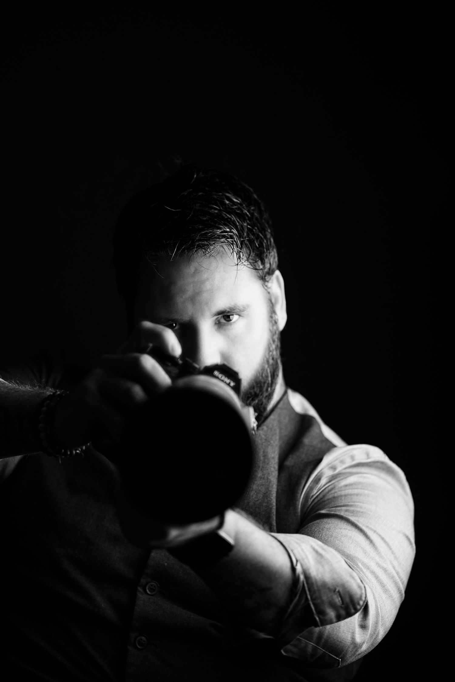 A photographer with a beard holds a professional camera in dramatic black and white lighting.